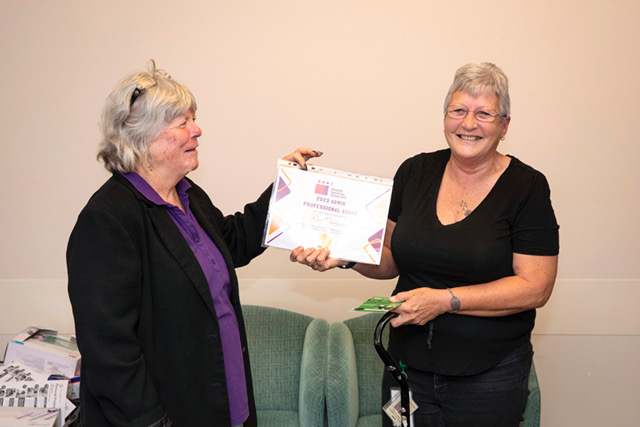HSWA Organiser Vickie McKay presents a certificate to Admin Professional Ray Cummins. Vickie wears a purple shirt and smiles at Rae who holds her certificate in one hand and smiles at the camera.