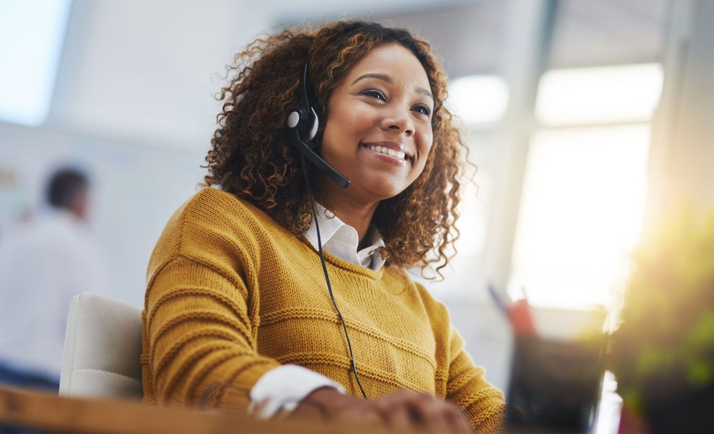 African woman with curly hair, and a yellow jumper smiles as she takes a call via her headset.