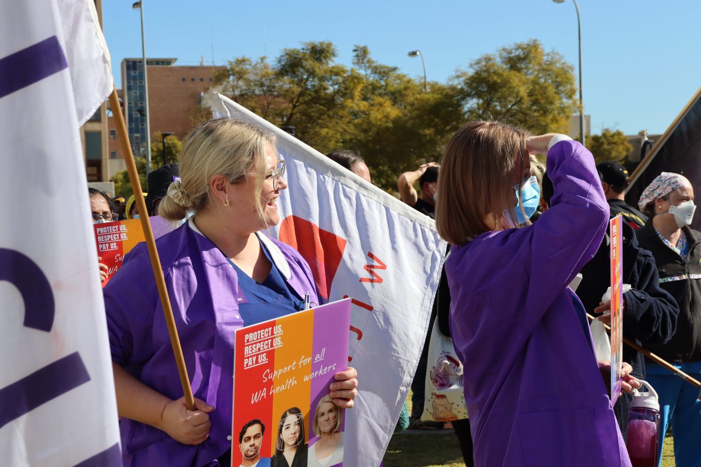 A young, blonde female health worker wearing glasses and a purple jacket over her scrubs, stands smiling and looking into the distance. She holds a HSUWA flag while standing in front of another. In her left hand she holds a HSUWA poster. She is standing in a crowd of people at a Stop Work Meeting at QEII Medical Campus.