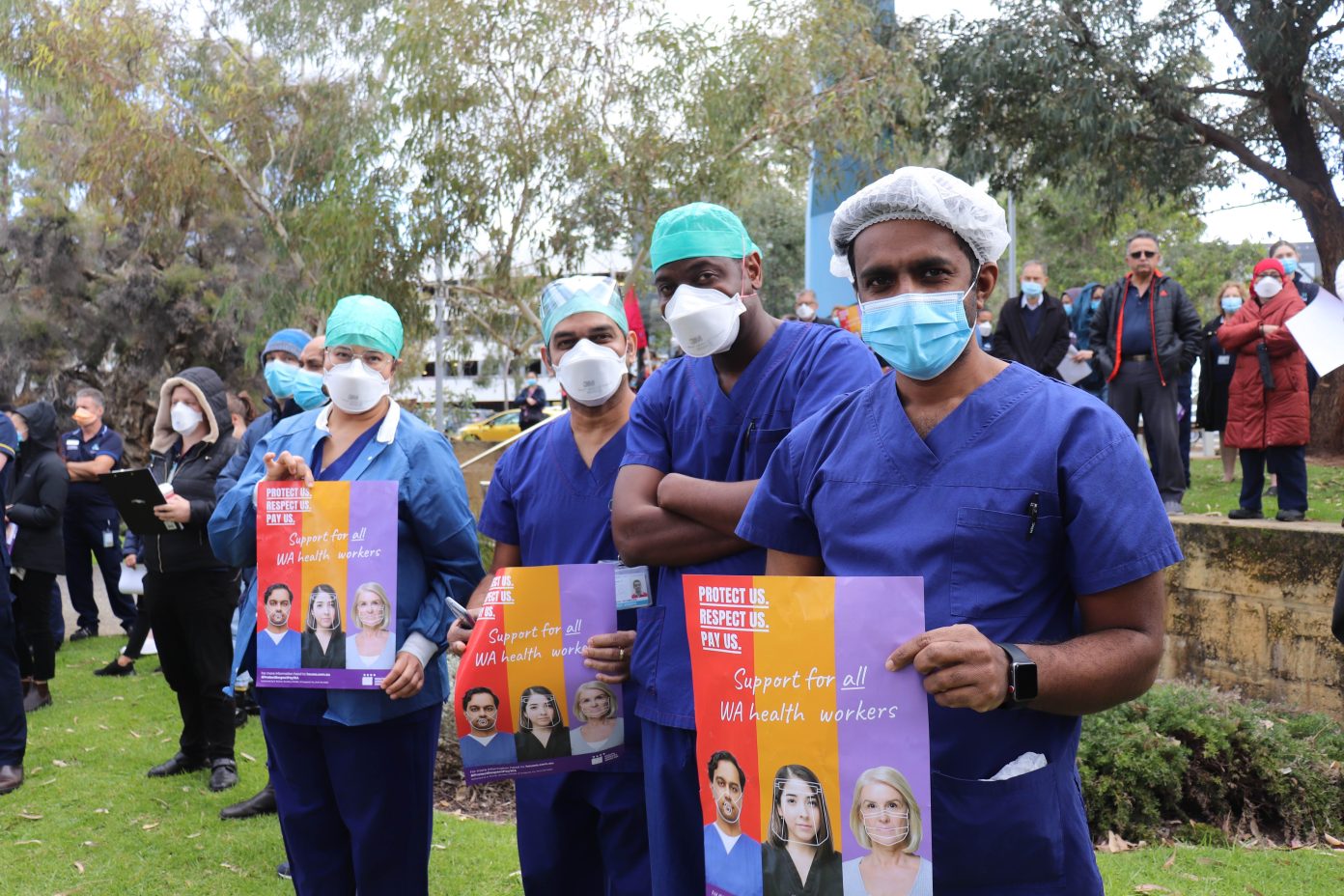 Four health workers wearing scrubs and masks stand outside Fiona Stanley hospital with HSUWA posters.
