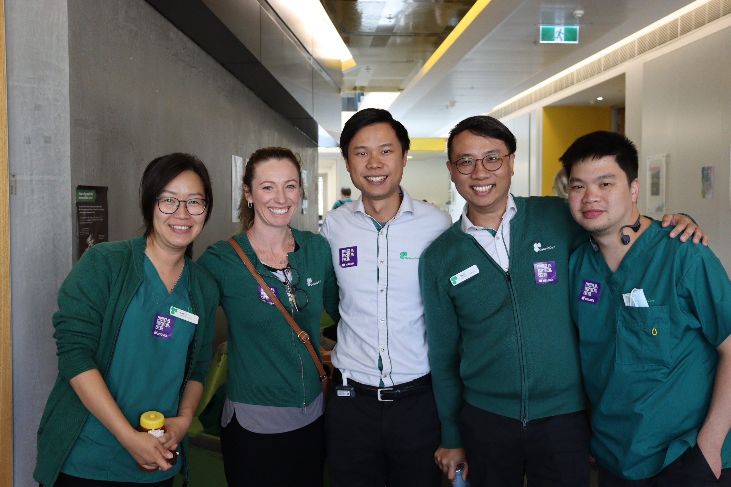 A group of young Radiation Therapists at Genesis Care stand smiling with their arms around each other. They are dressed professionally in their uniforms and wear purple union stickers.
