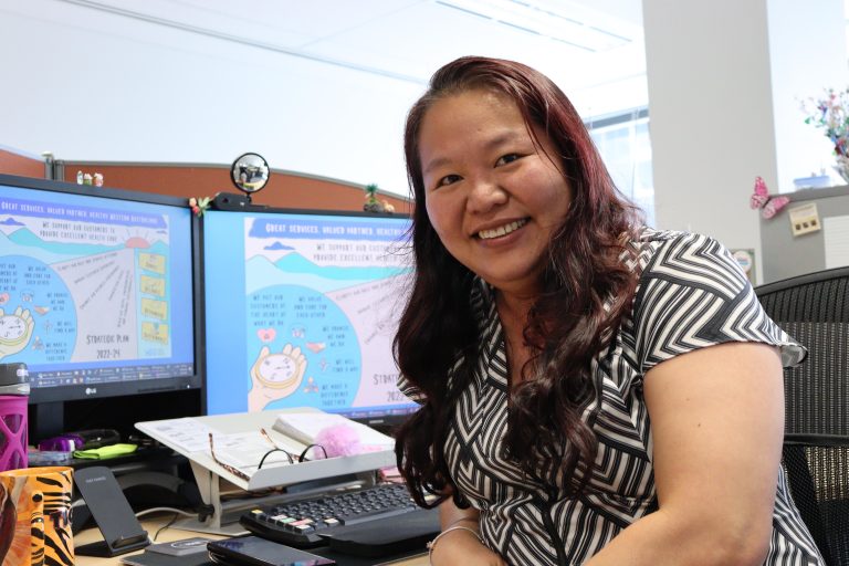 Female HSUWA Member sits at her desk at Health Support Services. She is smiling with two computer screens behind her.