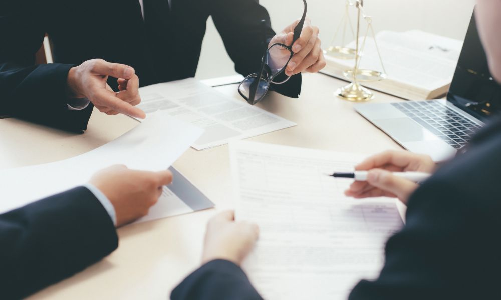 Three people sit around a table with legal paperwork. The image focuses on three pairs of hands with gold justice scales in the background.