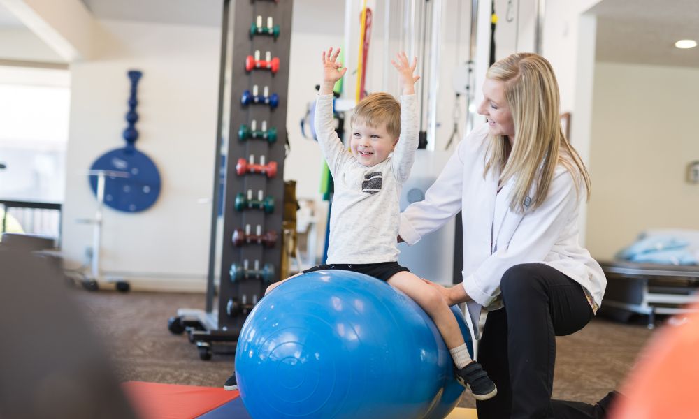 A female physiotherapist assists a small boy sitting on a fit ball, with his arms raised.