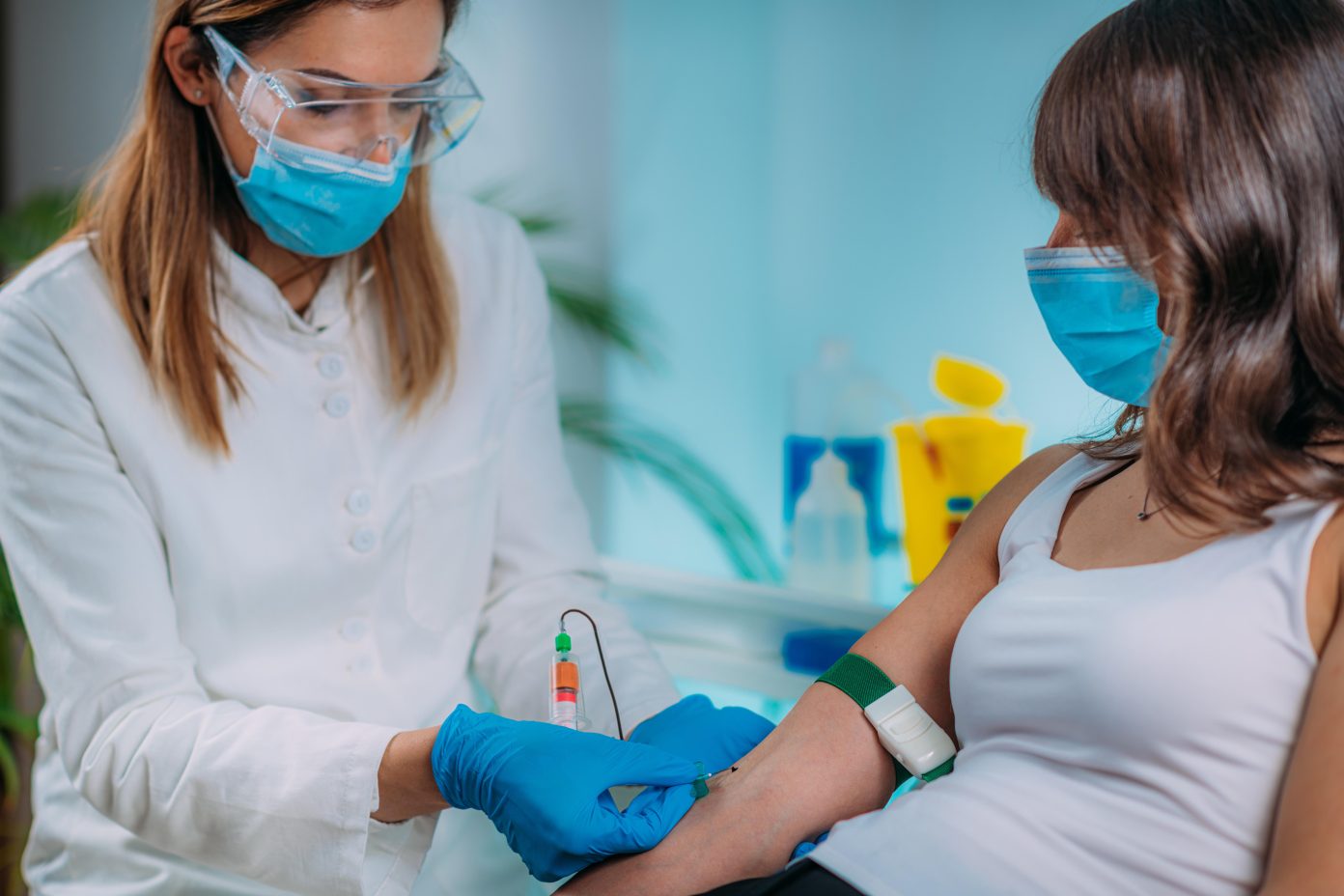 A female phlebotomist wearing safety glasses, a surgical mask and protective gloves prepares to collect a blood sample from a patient.