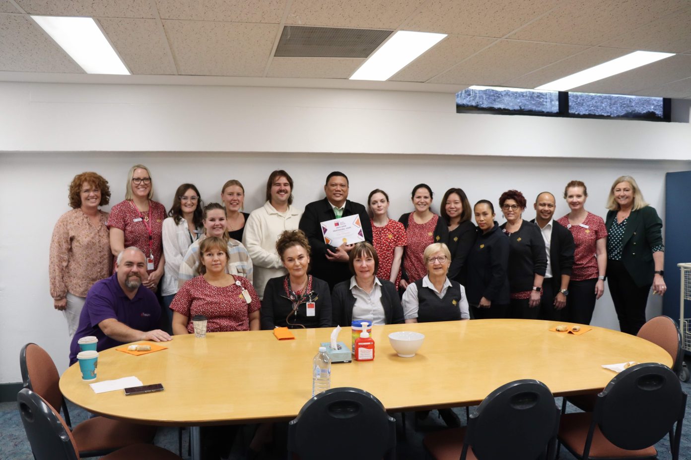 A group of health workers at St John of God Subiaco stand behind a lunch room table to celebrate their colleague who has won an award.