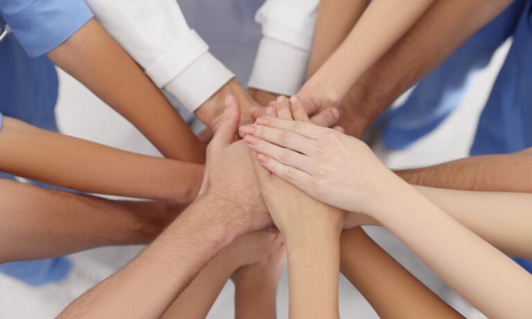 A close up of the hands of multiple health workers with various skin tones, stacking their hands together.