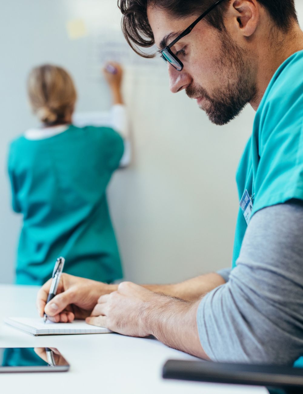 A male health worker wearing teal scrubs writes notes as his female colleague updates a roster in the background.