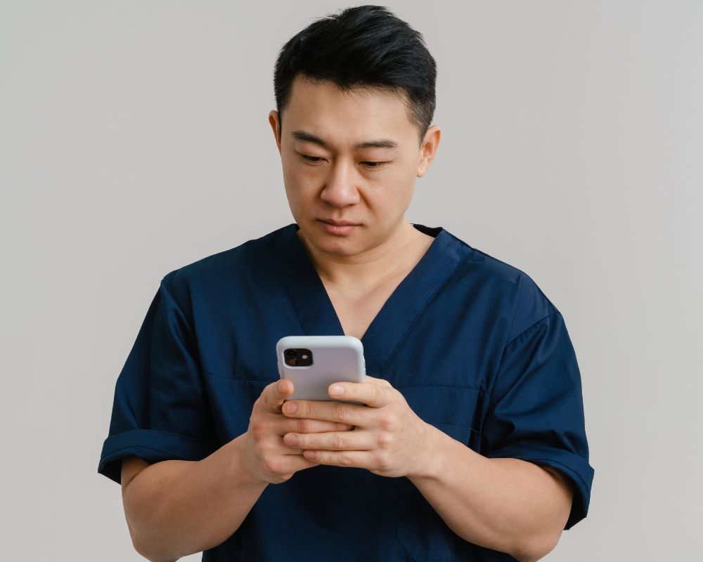 An Asian male health worker, wearing navy scrubs looks down at the smart phone he holds in both hands.