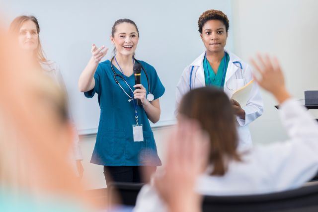 Health Professionals at a meeting facing an audience of other health professionals with their hands up.