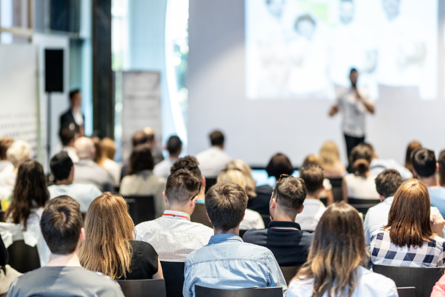 A large group of people dressed professionally attend a meeting. A presenter speaks at the front. The photograph is taken from the back of the room.