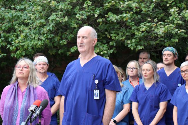 HSUWA Secretary Naomi McCrae and Anaesthetic Technician, Roy, speak to the media with Anaesthetic Technicians in blue scrubs standing behind them.