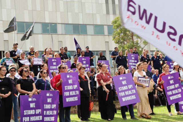 A group of health workers from Fiona Stanley Hospital stand together at a union rally holding flags and purple signs.