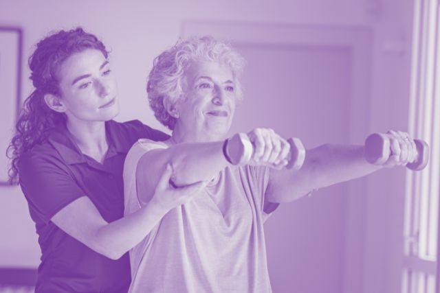 A female physiotherapist assists an elderly patient doing arm exercises with small dumbells.